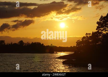 La photo capture un coucher de soleil sur un lac en Suède. Une forêt orne l'horizon, tandis que sur le côté droit, des formations rocheuses sont visibles. Ciel nuageux jaune Banque D'Images