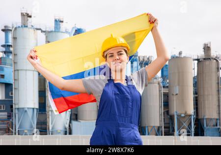 Positif jeune femme travailleuse dans le casque agitant le drapeau national de la Colombie tout en se tenant devant de grands réservoirs à l'usine de raffinerie le jour ensoleillé d'été Banque D'Images