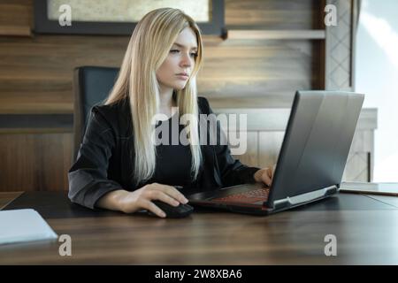 Fille blonde dans les vêtements d'affaires se trouve devant l'ordinateur portable dans le bureau et travaille intensément, concept d'éducation en ligne et de travail. Banque D'Images