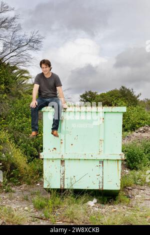 Craig Reucassel, comédien australien de la télévision et de la radio, connu pour l'équipe satirique The Chaser et The War on Waste. Assis sur la benne industrielle en métal. Banque D'Images