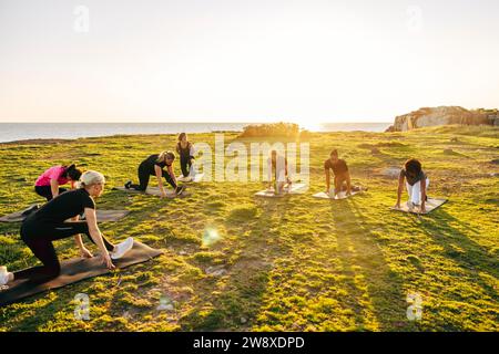 Équipe faisant des étirements sur l'herbe pendant la séance d'entraînement de groupe près de la mer contre le ciel Banque D'Images