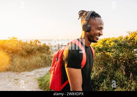 Jeune homme souriant portant un sac à dos et un casque sans fil contre le ciel Banque D'Images