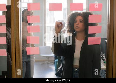 Femme d'affaires écrivant sur le mur de verre lors de la réunion avec des collègues au bureau Banque D'Images