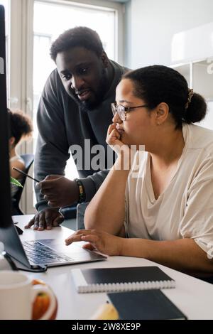 Collègue masculin discutant avec une collègue féminine assise avec la main sur le menton au bureau Banque D'Images