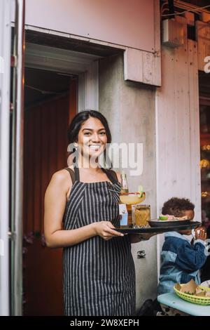 Portrait d'une serveuse souriante portant un tablier tenant un plateau de nourriture et de boissons au bar Banque D'Images