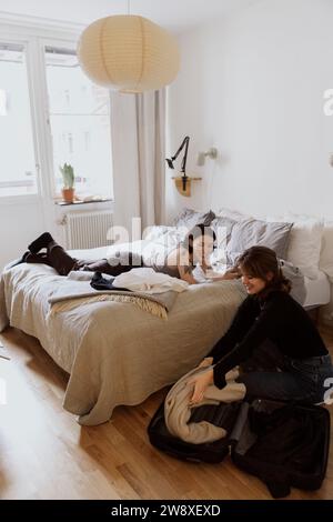 Femme souriante déballant les bagages près d'une amie couchée sur le lit dans l'appartement Banque D'Images