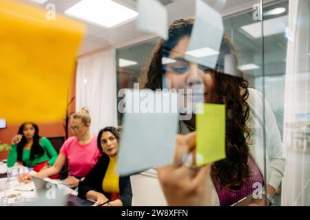 Jeune femme entrepreneure écrivant sur la note adhésive pendant la réunion d'affaires avec des collègues au bureau Banque D'Images