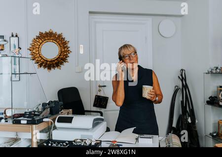 Vendeur senior avec tasse de café parlant sur le téléphone intelligent dans le magasin Banque D'Images