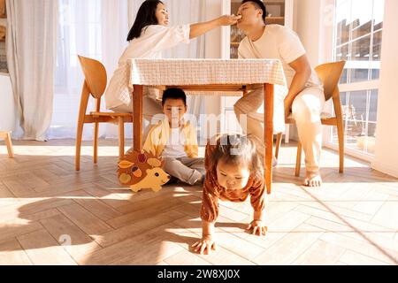 Les parents asiatiques prennent une collation à la maison dans la salle à manger. De mignons petits enfants jouent sous la table. Une famille coréenne heureuse s'amuse ensemble. Banque D'Images