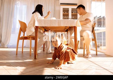 Les parents asiatiques prennent une collation à la maison dans la salle à manger. De mignons petits enfants jouent sous la table. Une famille coréenne heureuse s'amuse ensemble. Banque D'Images