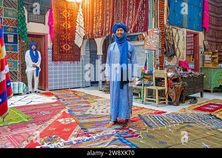 Vendeur marocain vendant des tapis et des tapis berbères tissés à la main dans un magasin de tapis dans la ville de Tinghir / Tinerhir, région de Drâa-Tafilalet, Maroc central Banque D'Images