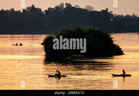 Silhouettes de personnes faisant du kayak à travers les eaux calmes du Mékong, dans l’archipel de si Phan Don, à travers des rayons de lumière dorée réfléchis par la riv Banque D'Images