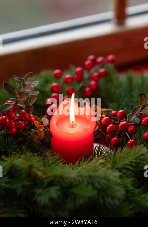 Couronne de l'Avent décorée de branches de sapin avec bougie allumée rouge sur un rebord de fenêtre en bois dans le temps avant Noël Banque D'Images