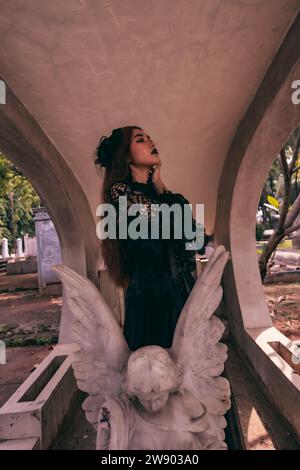 une jeune sorcière en robe noire se tient entre les pierres tombales en forme d'ange lors de la visite du cimetière dans l'après-midi Banque D'Images