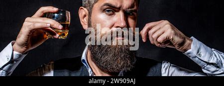 Un homme avec une barbe tient un verre de brandy. Concept dégustation et dégustation. Homme d'affaires barbu en costume élégant avec verre de whisky. Homme buvant du whisky Banque D'Images