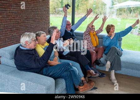 Cette image vivante capture un groupe de seniors entièrement immergés dans une compétition ludique de jeux vidéo. Assis sur un confortable canapé gris, ils présentent une gamme de réactions allant de l'attention concentrée au triomphe joyeux. Le cadre intérieur dispose d'une grande fenêtre avec vue sur l'extérieur verdoyant, offrant une atmosphère lumineuse et ouverte. Leurs expressions et gestes, des manettes serrées aux bras levés, reflètent l'excitation et le plaisir de l'activité partagée. Joyful Game Time, Seniors engagés dans la compétition ludique. Photo de haute qualité Banque D'Images