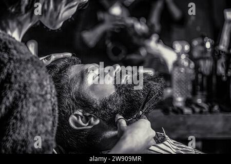 Barbier coupant les cheveux de l'homme barbu. Il se fait couper les cheveux au salon de coiffure. Salon de coiffure, salon de beauté. Homme de barbe dans le salon de coiffure. Ciseaux de barbier Banque D'Images