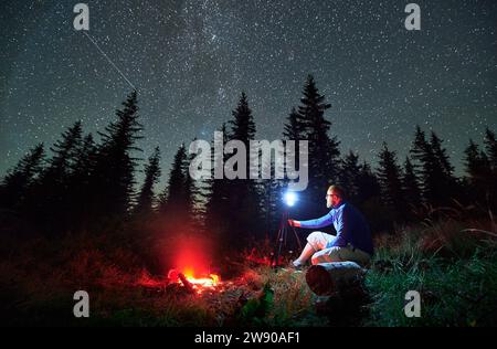 Photographe masculin faisant une séance photo dans les montagnes. Homme prenant des photos de nuit étoilée. Jeune homme assis sur un rondin à côté d'un feu de joie la nuit. Banque D'Images