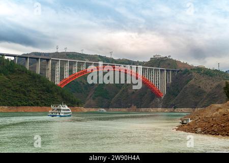 Une vue imprenable sur un pont rouge vibrant qui surplombe les eaux calmes du fleuve Yangtze en Chine, entouré de collines verdoyantes sous un ciel nuageux. Banque D'Images