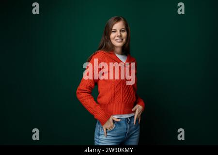 Belle femme mince aux cheveux bruns habillée d'un pull rouge et d'un Jean sur un fond vert Banque D'Images