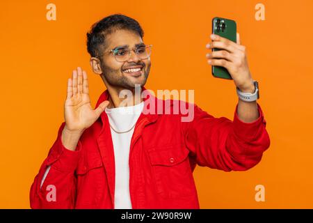 Souriant homme indien blogueur prenant le portrait selfie sur smartphone, streaming, vlog, communication d'enregistrement vidéo appel en ligne avec les abonnés de médias sociaux. Gars hindou arabe isolé sur fond orange Banque D'Images