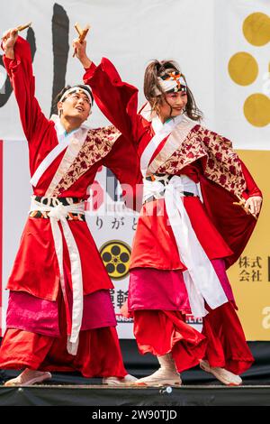 Gros plan, jeune femme et homme japonais yosakoi danseurs sur scène dansant en costume traditionnel rouge, avec un bras levé tenant naruko, claquettes. Banque D'Images
