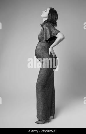Portrait noir et blanc de jeune jolie femme enceinte sur fond gris de studio. Femme en robe à sequins gris. Banque D'Images