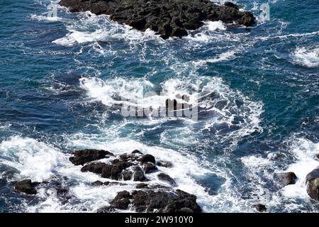 Mer bleue avec des vagues avec des chapeaux blancs. L'eau gonfle sauvagement et coule rapidement entre les roches noires dans l'eau Banque D'Images