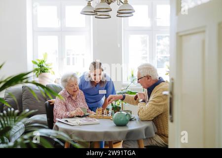 Travailleur de santé souriant avec un couple senior jouant aux échecs à table Banque D'Images