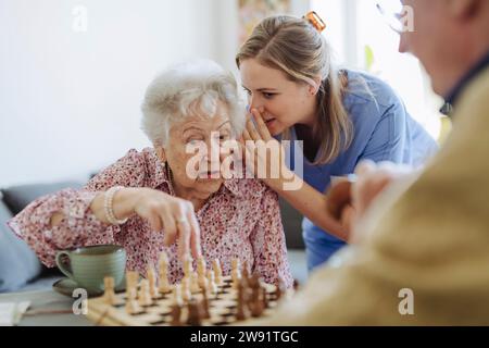 Travailleur de la santé chuchotant à l'oreille d'une femme âgée et jouant aux échecs à la maison Banque D'Images