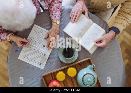 Homme senior lisant le livre avec la femme faisant le puzzle de mots croisés à la table Banque D'Images