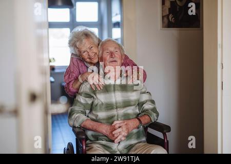 Heureuse femme âgée embrassant l'homme assis en fauteuil roulant à la maison Banque D'Images