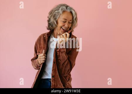 Femme âgée souriante portant une veste marron et debout sur fond rose Banque D'Images