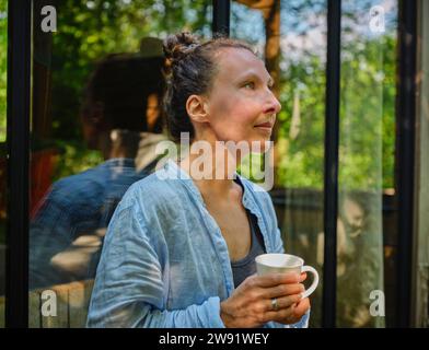 Femme mature tenant une tasse de café et se penchant sur la fenêtre Banque D'Images