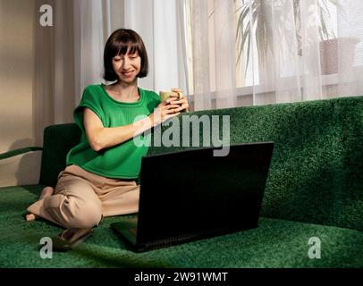 Souriant freelance tenant une tasse de thé et parlant sur appel vidéo via un ordinateur portable à la maison Banque D'Images