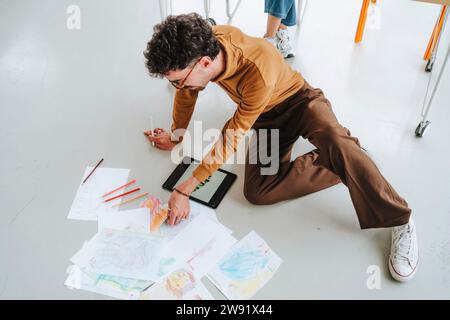 Jeune illustrateur examinant des dessins assis sur le sol au bureau Banque D'Images