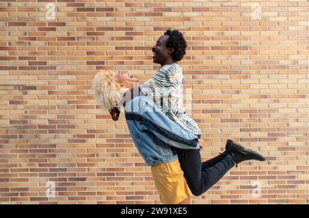 Femme transgenre joyeuse soulevant petit ami devant le mur de briques Banque D'Images