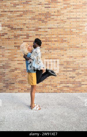 Femme transgenre soulevant son petit ami debout devant un mur de briques Banque D'Images