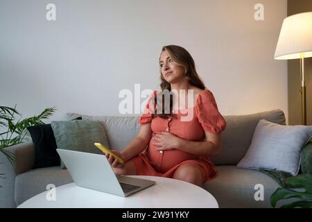 Femme enceinte réfléchie tenant un téléphone intelligent avec ordinateur portable sur la table à la maison Banque D'Images