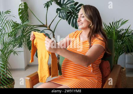 Femme enceinte souriante tenant des vêtements de bébé assis sur un fauteuil à la maison Banque D'Images