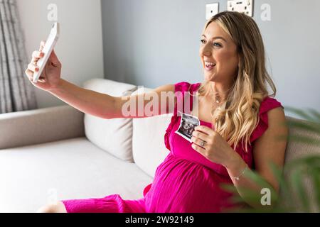 Femme enceinte souriante tenant la photographie d'échographie et prenant selfie par téléphone intelligent à la maison Banque D'Images