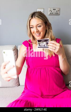 Femme enceinte souriante montrant la photographie d'échographie sur l'appel vidéo par téléphone intelligent à la maison Banque D'Images