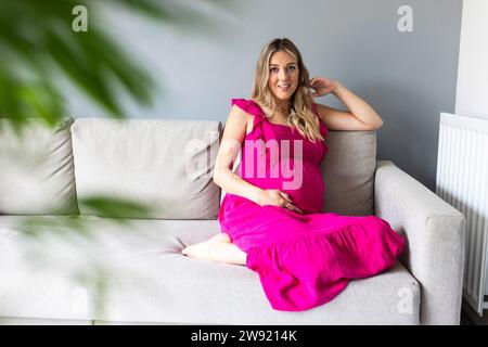 Femme enceinte souriante portant une robe rose et assise sur un canapé à la maison Banque D'Images