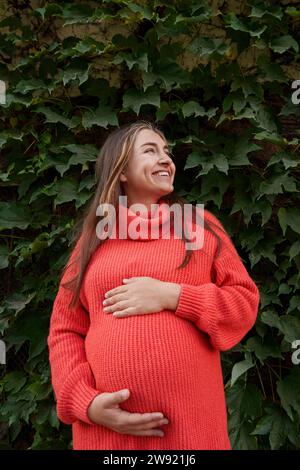 Femme enceinte souriante portant une robe rouge et debout devant les plantes Banque D'Images