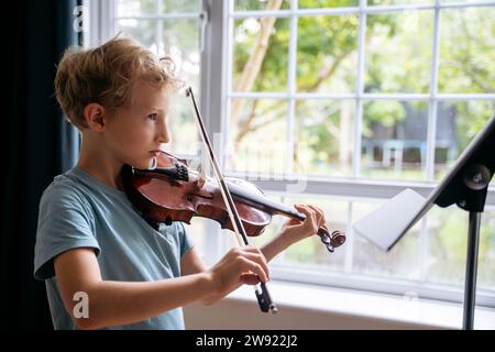 Garçon pratiquant le violon avec partition près de la fenêtre à la maison Banque D'Images