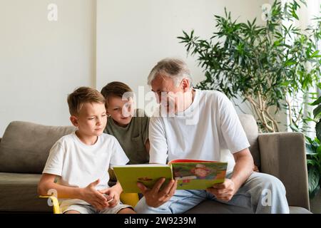 Homme âgé souriant lisant le livre pour les petits-fils dans le salon à la maison Banque D'Images