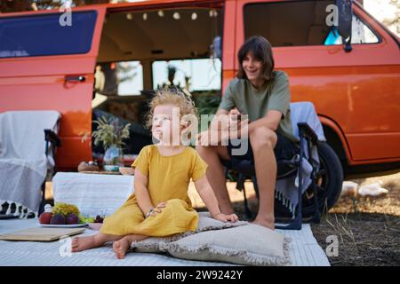 Fille assise sur la couverture de pique-nique avec frère sur la chaise par caravane Banque D'Images