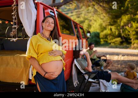 Femme enceinte souriante appuyée sur la caravane par la famille assise dans le parc Banque D'Images
