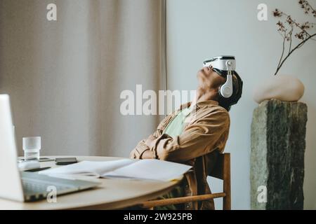 Jeune homme portant un casque VR assis avec les bras croisés et la tête en arrière à la table Banque D'Images