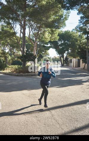 Femme de fitness mature, entre 30 et 50 ans, courant, sur la route, entouré de pins, portant des lunettes de sport, casque bluetooth, bleu Banque D'Images